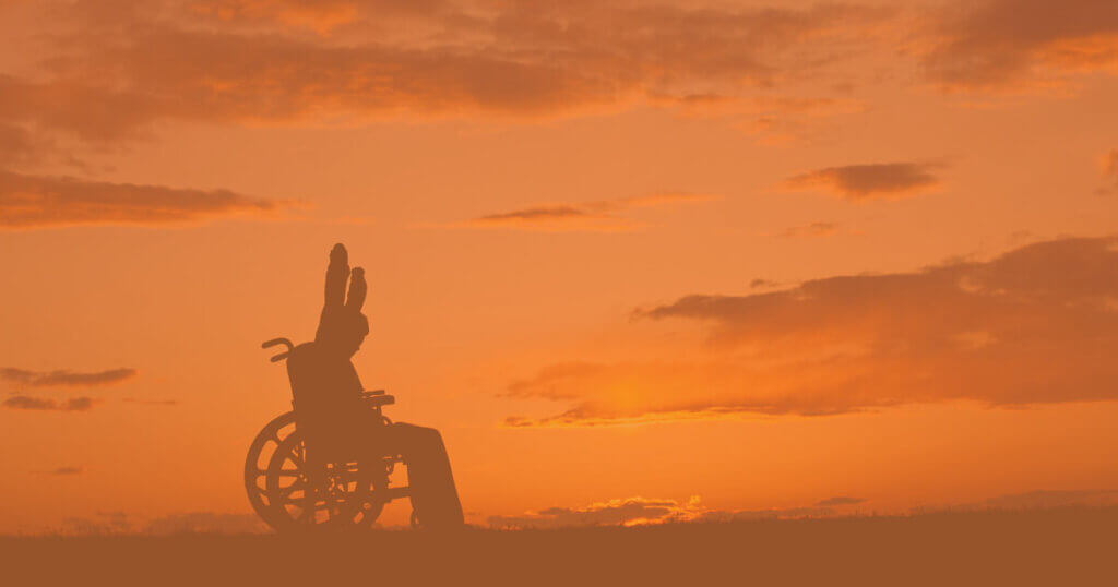 person in a wheelchair throws their arms up in trumph behind a setting sun.